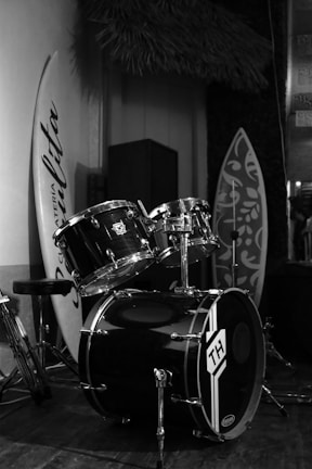 A black drum set is positioned in an indoor setting, surrounded by surfboards. The drums display a logo with the letters 'TH', and the surrounding area appears decorated with textured walls and plants, suggesting a casual or beach-themed ambiance.