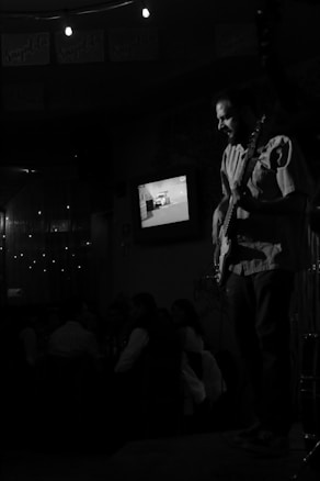 A live music performance in a dimly lit venue features a guitarist playing on stage while people sit at tables in the background. A television screen on the wall displays an image, and string lights add to the ambiance.