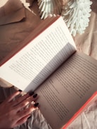 An open book held by a hand with dark nail polish, surrounded by brown textured paper and small white decorative objects resembling trees. Sunlight casts shadows across the book's pages.