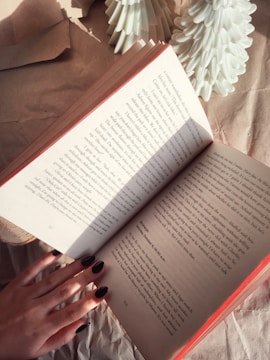 An open book held by a hand with dark nail polish, surrounded by brown textured paper and small white decorative objects resembling trees. Sunlight casts shadows across the book's pages.