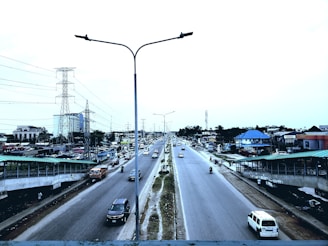 A busy urban road with multiple vehicles, including cars and motorcycles, moving in both directions. Streetlights are positioned along the median, and there are pedestrian walkways on either side of the road. Buildings of various sizes, including residential and commercial structures, line the sides of the road. Power lines and a transmission tower are visible in the background, indicating infrastructure development.