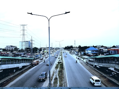 A busy urban road with multiple vehicles, including cars and motorcycles, moving in both directions. Streetlights are positioned along the median, and there are pedestrian walkways on either side of the road. Buildings of various sizes, including residential and commercial structures, line the sides of the road. Power lines and a transmission tower are visible in the background, indicating infrastructure development.