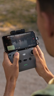 A person is holding a DJI remote controller with a smartphone attached. The phone screen displays a live video feed of a landscape, possibly from a drone, with trees and water visible. The person's hands are gripping the controller while standing outdoors.