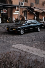 A classic dark-colored car is parked on a cobblestone street in front of a building with a neon sign reading 'FOOD INDUSTRY'. The building features large windows decorated with fairy lights and has an industrial vibe with visible air conditioning units. There are plants and architectural details like an awning and signage that contribute to a vintage urban atmosphere.