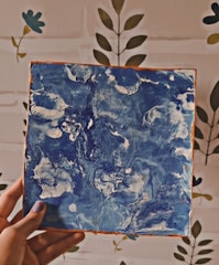 A hand is holding a square canvas with abstract blue and white swirled patterns. The background features a wall with a simple floral design.