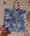 A hand is holding a square canvas with abstract blue and white swirled patterns. The background features a wall with a simple floral design.