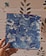 A hand is holding a square canvas with abstract blue and white swirled patterns. The background features a wall with a simple floral design.