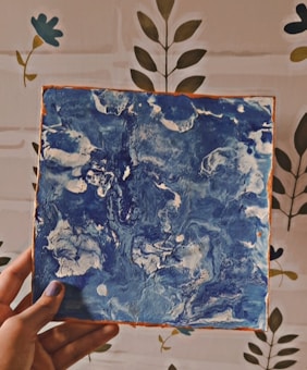 A hand is holding a square canvas with abstract blue and white swirled patterns. The background features a wall with a simple floral design.