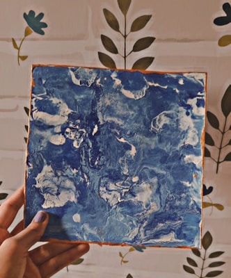 A hand is holding a square canvas with abstract blue and white swirled patterns. The background features a wall with a simple floral design.