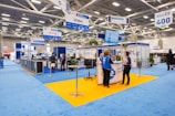 An indoor exhibition hall featuring various booths with blue signage and carpeting. People are conversing and navigating the space, which is brightly lit by overhead lighting. Portable signs and technology displays are visible throughout, and the space is open with a modern design.