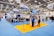 An indoor exhibition hall featuring various booths with blue signage and carpeting. People are conversing and navigating the space, which is brightly lit by overhead lighting. Portable signs and technology displays are visible throughout, and the space is open with a modern design.