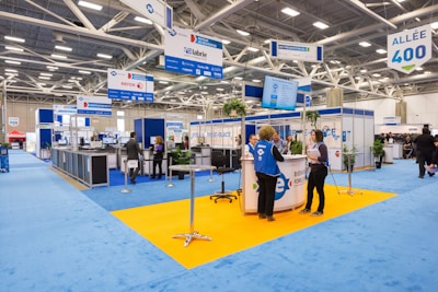 An indoor exhibition hall featuring various booths with blue signage and carpeting. People are conversing and navigating the space, which is brightly lit by overhead lighting. Portable signs and technology displays are visible throughout, and the space is open with a modern design.