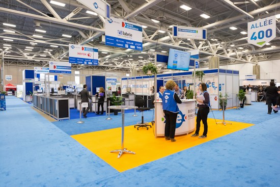 An indoor exhibition hall featuring various booths with blue signage and carpeting. People are conversing and navigating the space, which is brightly lit by overhead lighting. Portable signs and technology displays are visible throughout, and the space is open with a modern design.
