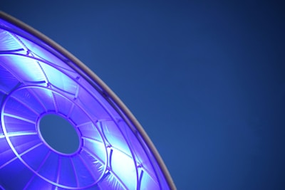 A circular, geometric structure glows with a bright blue light against a deep blue background. The structure appears to be part of a larger framework, with visible spokes or beams radiating from a central circular opening.