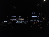 Neon signs of various restaurants and businesses are illuminated against a dimly lit background. The signs include names like Harjalis Restaurant, Octant Pizza, and Madhuram Hall, with letters glowing in bright white or varying colors. The overall scene is dark, highlighting the vibrant signage.