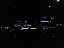 Neon signs of various restaurants and businesses are illuminated against a dimly lit background. The signs include names like Harjalis Restaurant, Octant Pizza, and Madhuram Hall, with letters glowing in bright white or varying colors. The overall scene is dark, highlighting the vibrant signage.