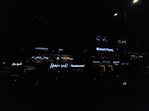 Neon signs of various restaurants and businesses are illuminated against a dimly lit background. The signs include names like Harjalis Restaurant, Octant Pizza, and Madhuram Hall, with letters glowing in bright white or varying colors. The overall scene is dark, highlighting the vibrant signage.