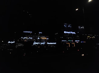 Neon signs of various restaurants and businesses are illuminated against a dimly lit background. The signs include names like Harjalis Restaurant, Octant Pizza, and Madhuram Hall, with letters glowing in bright white or varying colors. The overall scene is dark, highlighting the vibrant signage.
