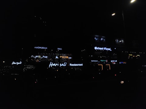 Neon signs of various restaurants and businesses are illuminated against a dimly lit background. The signs include names like Harjalis Restaurant, Octant Pizza, and Madhuram Hall, with letters glowing in bright white or varying colors. The overall scene is dark, highlighting the vibrant signage.