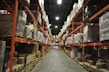 A large warehouse with high shelves stacked with boxes and packages. The shelves are metallic and color-coded in orange. The floor is wide and clear, allowing movement for forklifts or workers. The ceiling is high with industrial lighting.