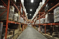 A large warehouse with high shelves stacked with boxes and packages. The shelves are metallic and color-coded in orange. The floor is wide and clear, allowing movement for forklifts or workers. The ceiling is high with industrial lighting.