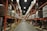A large warehouse with high shelves stacked with boxes and packages. The shelves are metallic and color-coded in orange. The floor is wide and clear, allowing movement for forklifts or workers. The ceiling is high with industrial lighting.