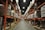 A large warehouse with high shelves stacked with boxes and packages. The shelves are metallic and color-coded in orange. The floor is wide and clear, allowing movement for forklifts or workers. The ceiling is high with industrial lighting.