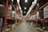 A large warehouse with high shelves stacked with boxes and packages. The shelves are metallic and color-coded in orange. The floor is wide and clear, allowing movement for forklifts or workers. The ceiling is high with industrial lighting.