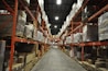 A large warehouse with high shelves stacked with boxes and packages. The shelves are metallic and color-coded in orange. The floor is wide and clear, allowing movement for forklifts or workers. The ceiling is high with industrial lighting.