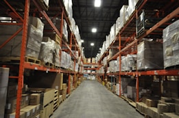 A large warehouse with high shelves stacked with boxes and packages. The shelves are metallic and color-coded in orange. The floor is wide and clear, allowing movement for forklifts or workers. The ceiling is high with industrial lighting.