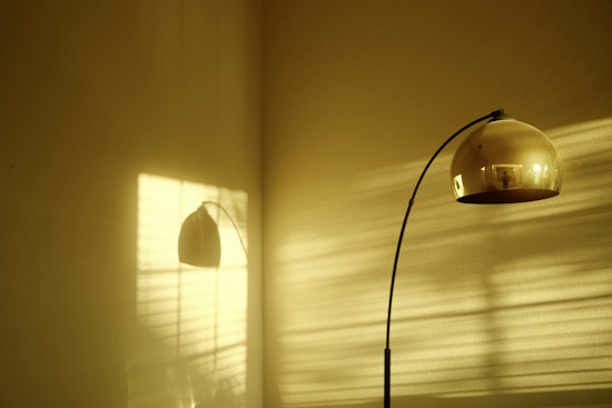 A minimalistic scene featuring a modern, curved floor lamp with a large, spherical gold shade. The lamp casts a shadow on a plain wall with a warm, golden hue, while horizontal shadows from a window blind create a striped pattern across the surface.