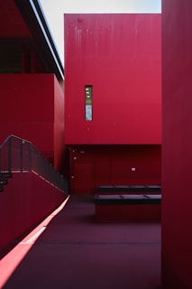An architectural scene featuring bold, red geometric structures with sharp lines and shadows. The design includes a tall red wall with a narrow window, a staircase, and benches or platforms, creating a minimalist and modern aesthetic.