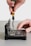 A hand is using a screwdriver to work on an open electronic device, possibly a hard drive or similar component. The device is positioned on a flat surface, and intricate parts and circuit boards are visible inside.