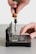 A hand is using a screwdriver to work on an open electronic device, possibly a hard drive or similar component. The device is positioned on a flat surface, and intricate parts and circuit boards are visible inside.