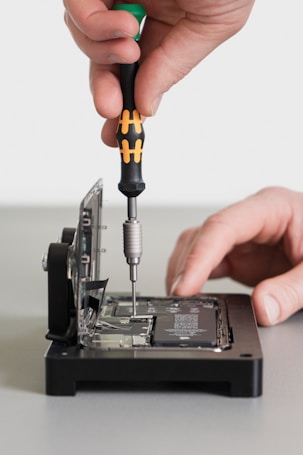A hand is using a screwdriver to work on an open electronic device, possibly a hard drive or similar component. The device is positioned on a flat surface, and intricate parts and circuit boards are visible inside.