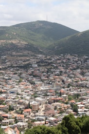 A sprawling town at the base of lush green hills. Numerous buildings with light-colored walls and red roofs are densely packed together. The landscape includes a mix of residential and possibly commercial structures, with winding streets and scattered vegetation.