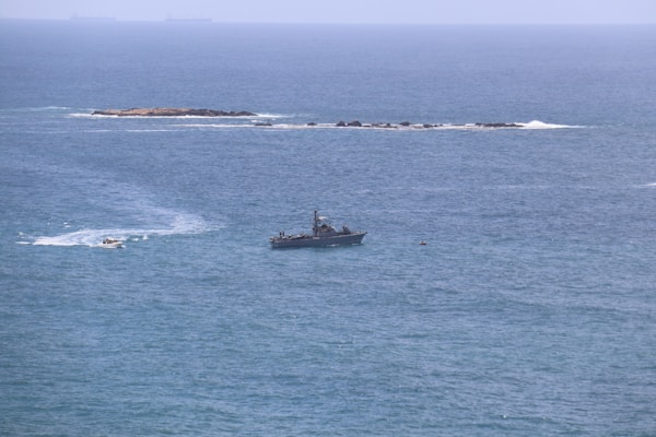 A military ship is positioned on the open sea, with an island and distant cargo ships in the background. The water is calm, and a smaller boat can be seen making waves near the ship.