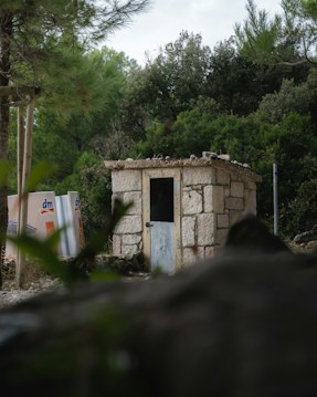 A small stone building with a simple door is nestled among lush green trees and foliage. Two white containers with 'dm' logos are visible nearby, adding a contrast to the natural surroundings. A rocky foreground frames the view, enhancing the secluded and rural atmosphere.