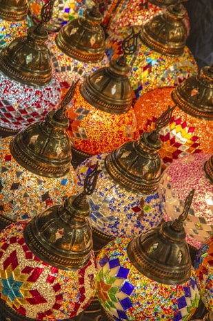 A collection of intricately designed, colorful mosaic lamps featuring geometric patterns in vibrant colors. The lamps have ornate, metallic tops and are tightly arranged, creating a rich tapestry of colors and textures.
