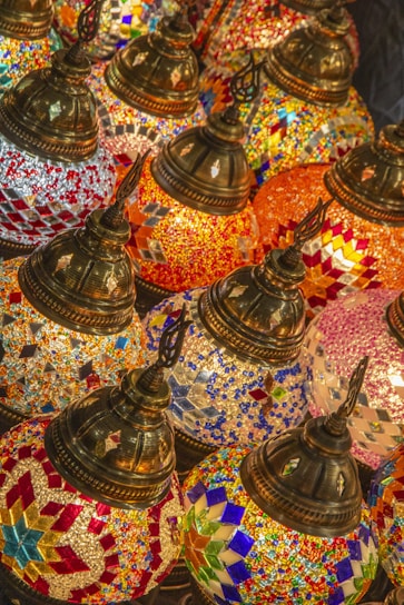 A collection of intricately designed, colorful mosaic lamps featuring geometric patterns in vibrant colors. The lamps have ornate, metallic tops and are tightly arranged, creating a rich tapestry of colors and textures.