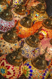 A collection of intricately designed, colorful mosaic lamps featuring geometric patterns in vibrant colors. The lamps have ornate, metallic tops and are tightly arranged, creating a rich tapestry of colors and textures.