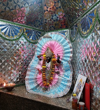A richly adorned idol surrounded by vibrant cloth and intricate patterns. The background consists of reflective mosaic tiles in various colors, creating a shimmering effect. An oil lamp flickers on a granite surface, alongside some offerings and religious art, contributing to a sacred atmosphere.