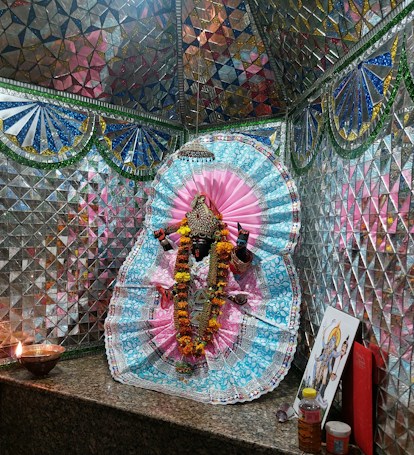 A richly adorned idol surrounded by vibrant cloth and intricate patterns. The background consists of reflective mosaic tiles in various colors, creating a shimmering effect. An oil lamp flickers on a granite surface, alongside some offerings and religious art, contributing to a sacred atmosphere.