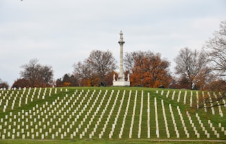 Rows of white headstones are neatly arranged in a cemetery, set against a backdrop of autumn trees with orange and brown foliage. A prominent stone monument topped with a statue is visible in the center, standing on a small hill.
