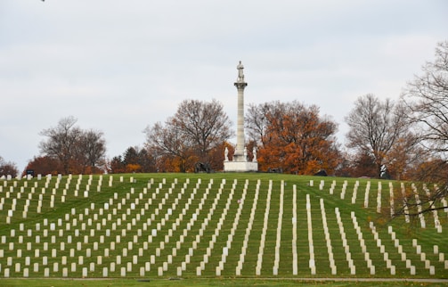Rows of white headstones are neatly arranged in a cemetery, set against a backdrop of autumn trees with orange and brown foliage. A prominent stone monument topped with a statue is visible in the center, standing on a small hill.