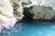 A natural cave formation with rocky walls and an opening at the far end. The cave is partially filled with a calm turquoise water, and light illuminates parts of the cave's interior, revealing details of the rock texture and some patches of moss.