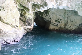 A natural cave formation with rocky walls and an opening at the far end. The cave is partially filled with a calm turquoise water, and light illuminates parts of the cave's interior, revealing details of the rock texture and some patches of moss.