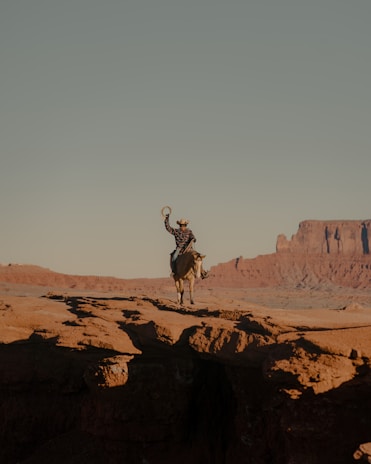 A person riding a horse stands on the edge of a canyon in a desert landscape, with the rider holding up a hat. The expansive scene includes rocky formations and a wide, clear sky.
