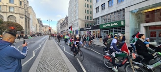 A group of cyclists rides through a city street, participating in what appears to be a demonstration or parade. Several riders hold flags, including one with a recognizable red, black, white, and green pattern. The city street is lined with various commercial buildings, including a Lloyds Bank. People are seen walking along the sidewalks, and one individual in the foreground is using a phone to capture the scene.