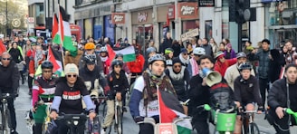 A group of cyclists participating in a protest ride through a city street, carrying Palestinian flags and wearing protest-themed clothing. The crowd is diverse and exhibits solidarity, with some participants holding signs. The street is lined with buildings and storefronts, filled with spectators and more demonstrators.